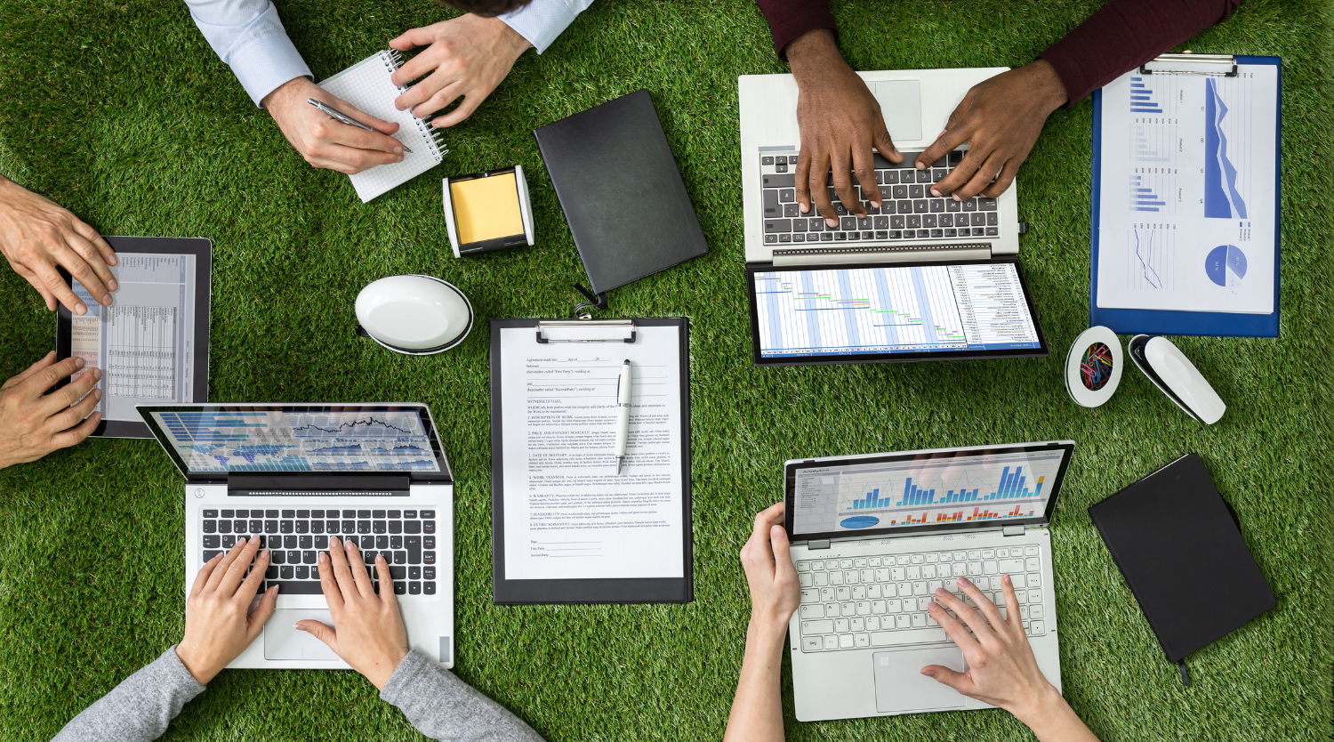 Weblio team collaborating outdoors with laptops and documents on green grass